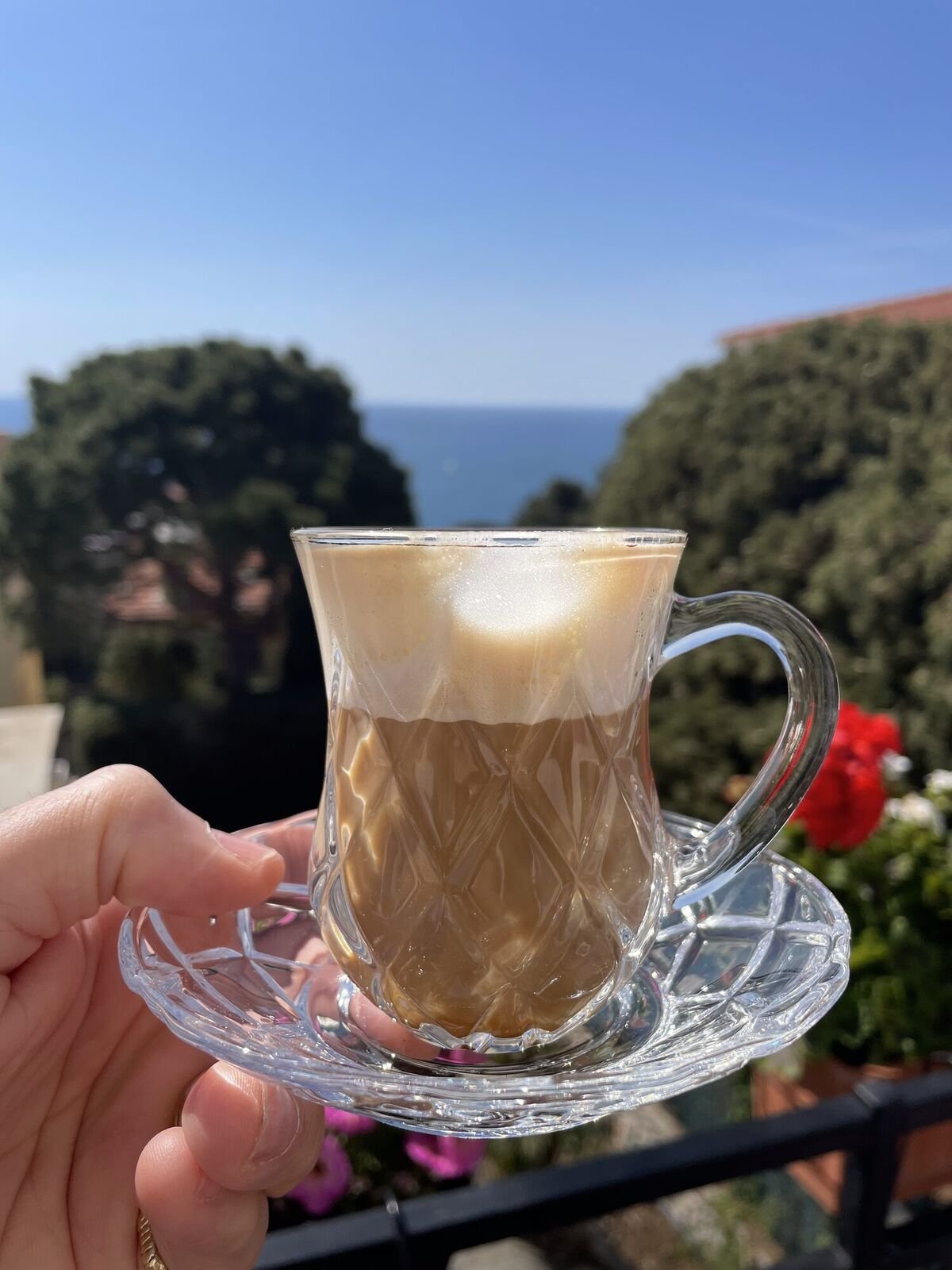 A glass of café crème held up against the sea on the terrace